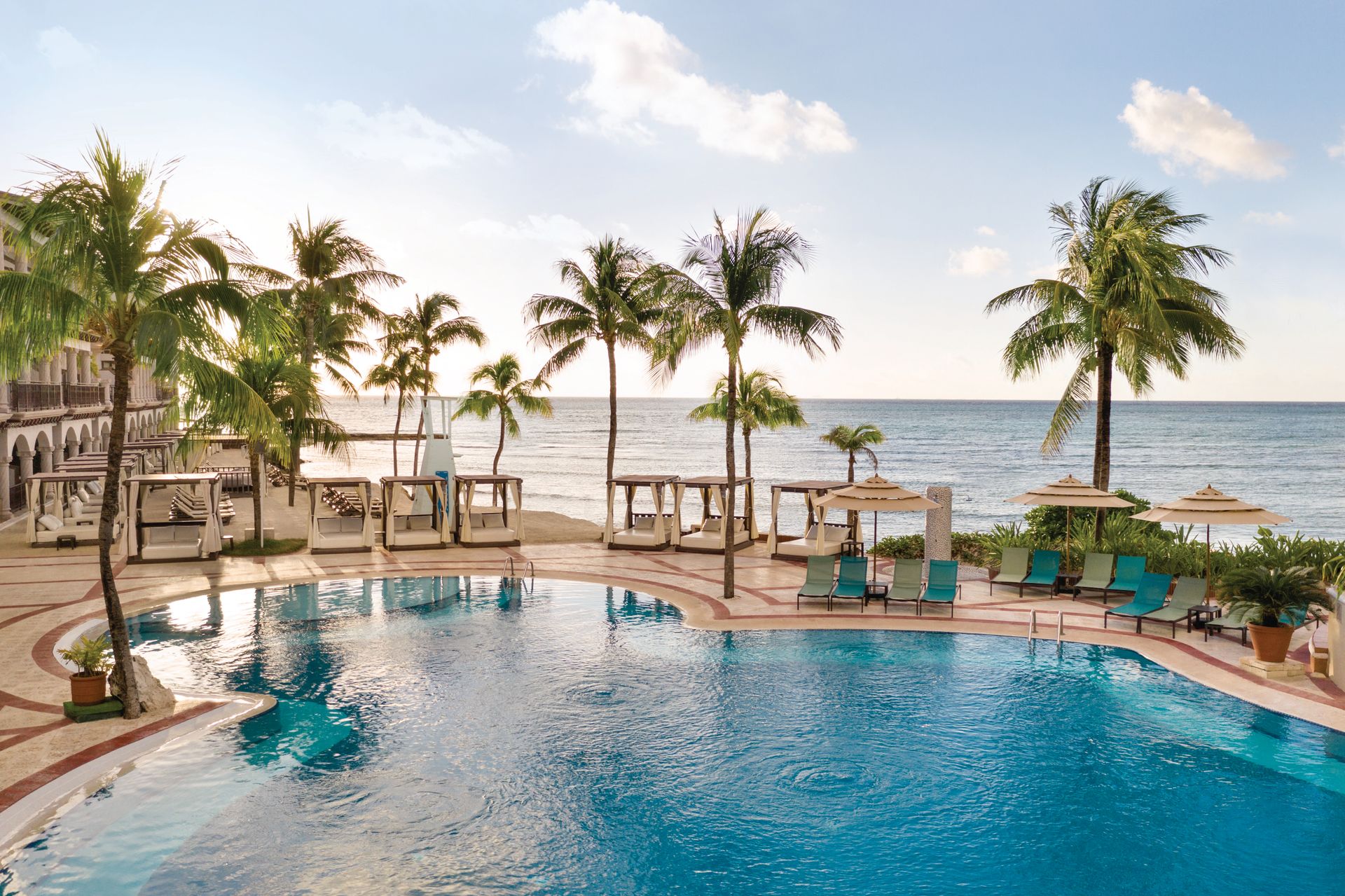 Beachfront infinity pool with turquoise water, palm trees, thatched umbrellas, and lounge chairs overlooking the Caribbean Sea at Hilton Playa del Carmen all-inclusive resort
