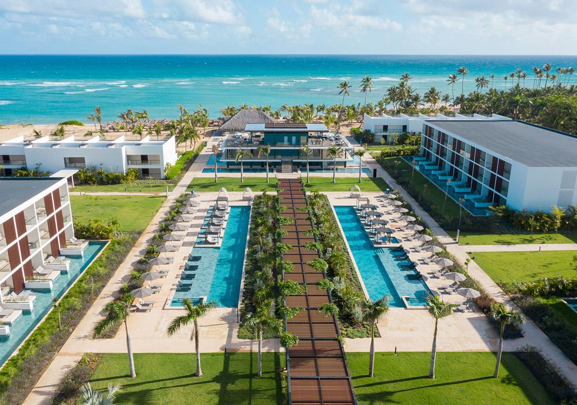 Aerial view of a modern beachfront resort with long pools, lounge chairs, palm trees, and the turquoise ocean stretching beyond the property.