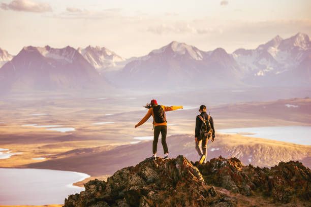 Two hikers stand on a rocky mountain summit overlooking a vast landscape of lakes, valleys, and snow-capped peaks at sunrise. The breathtaking view captures the freedom, adventure, and unforgettable memories that come from exploring the world. Two hikers stand on a rocky mountain summit overlooking a vast landscape of lakes, valleys, and snow-capped peaks at sunrise. The breathtaking view captures the freedom, adventure, and unforgettable memories that come from exploring the world.