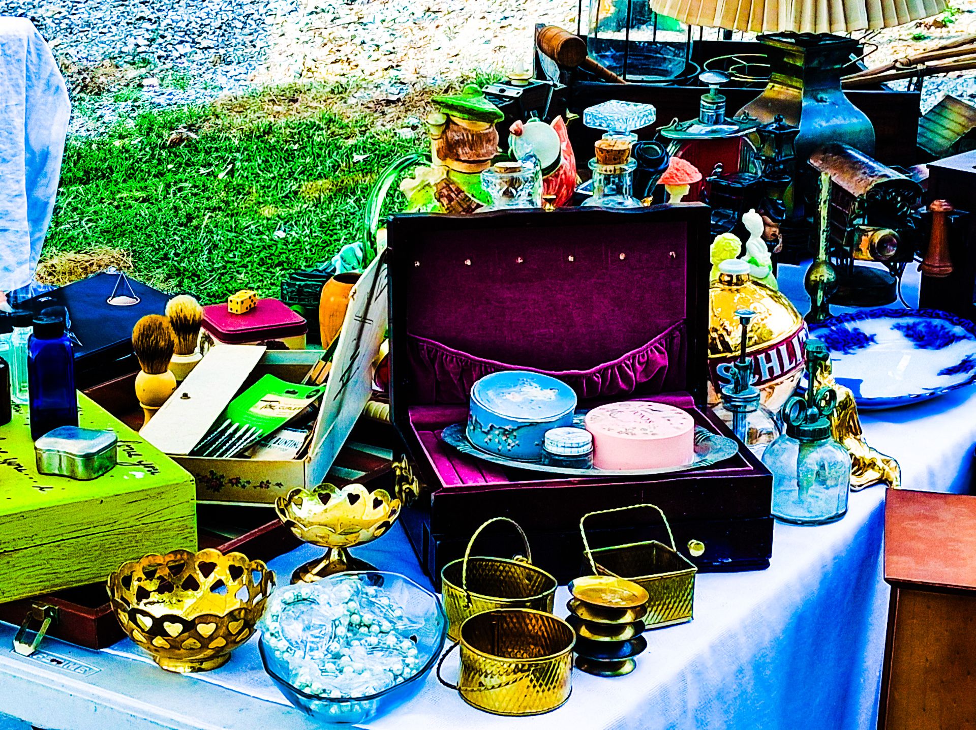 “Colorful display of assorted vintage items at a garage sale, including brass bowls, shaving brushes, glass bottles, and a velvet-lined case with trinkets.”