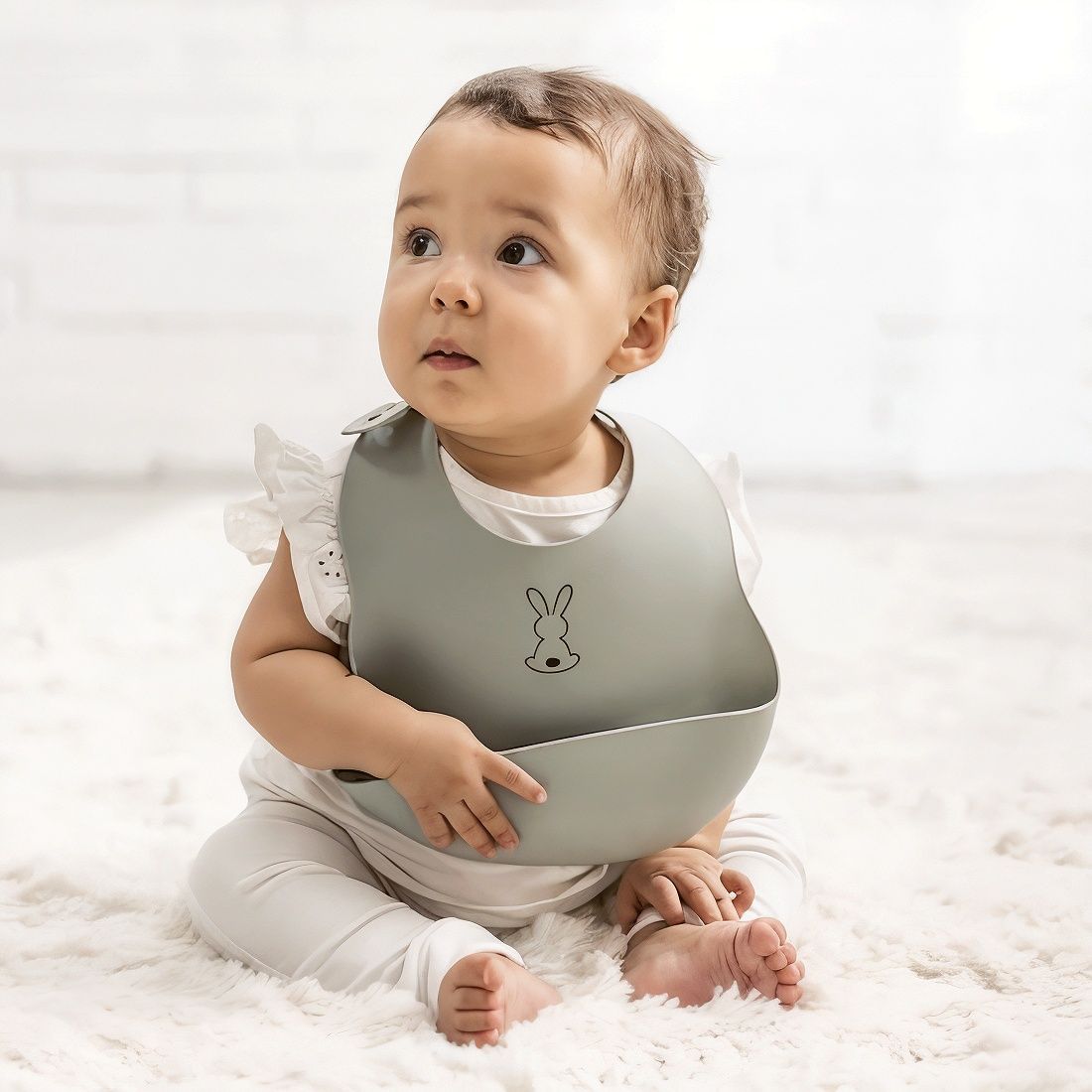 bébé portant un bavoir, assis sur un tapis blanc, regardant quelque chose d'un air inquiet. bébé portant un bavoir, assis sur un tapis blanc, regardant quelque chose d'un air inquiet.