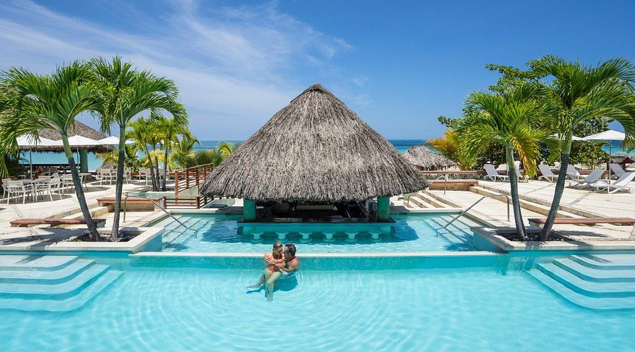 Couple relaxing together in a tropical resort infinity pool in Negril, Jamaica with a swim-up bar, palm trees, and ocean views under a bright blue sky.