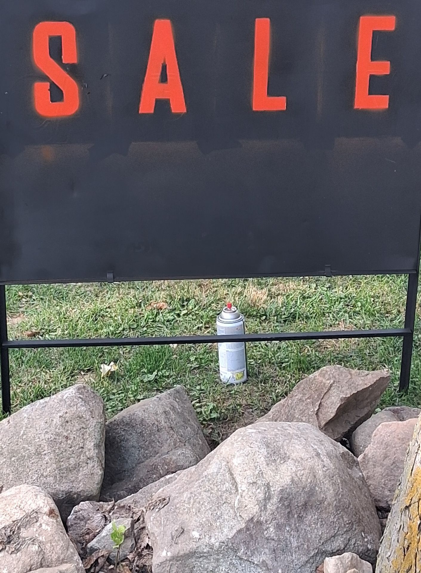 Black outdoor sign with bold red stenciled letters spelling SALE and a spray paint can on the grass in front of rocks.