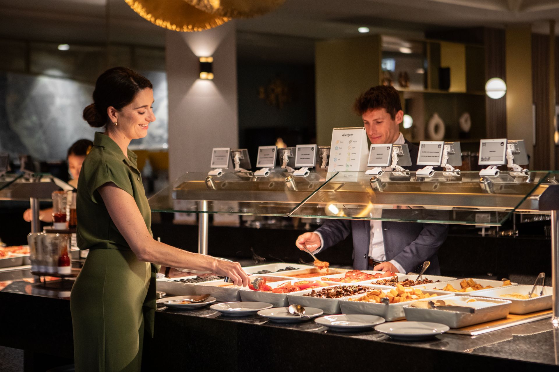 Guests serving themselves at an elegant all-inclusive resort buffet with a variety of dishes under warm lighting. Guests serving themselves at an elegant all-inclusive resort buffet with a variety of dishes under warm lighting.
