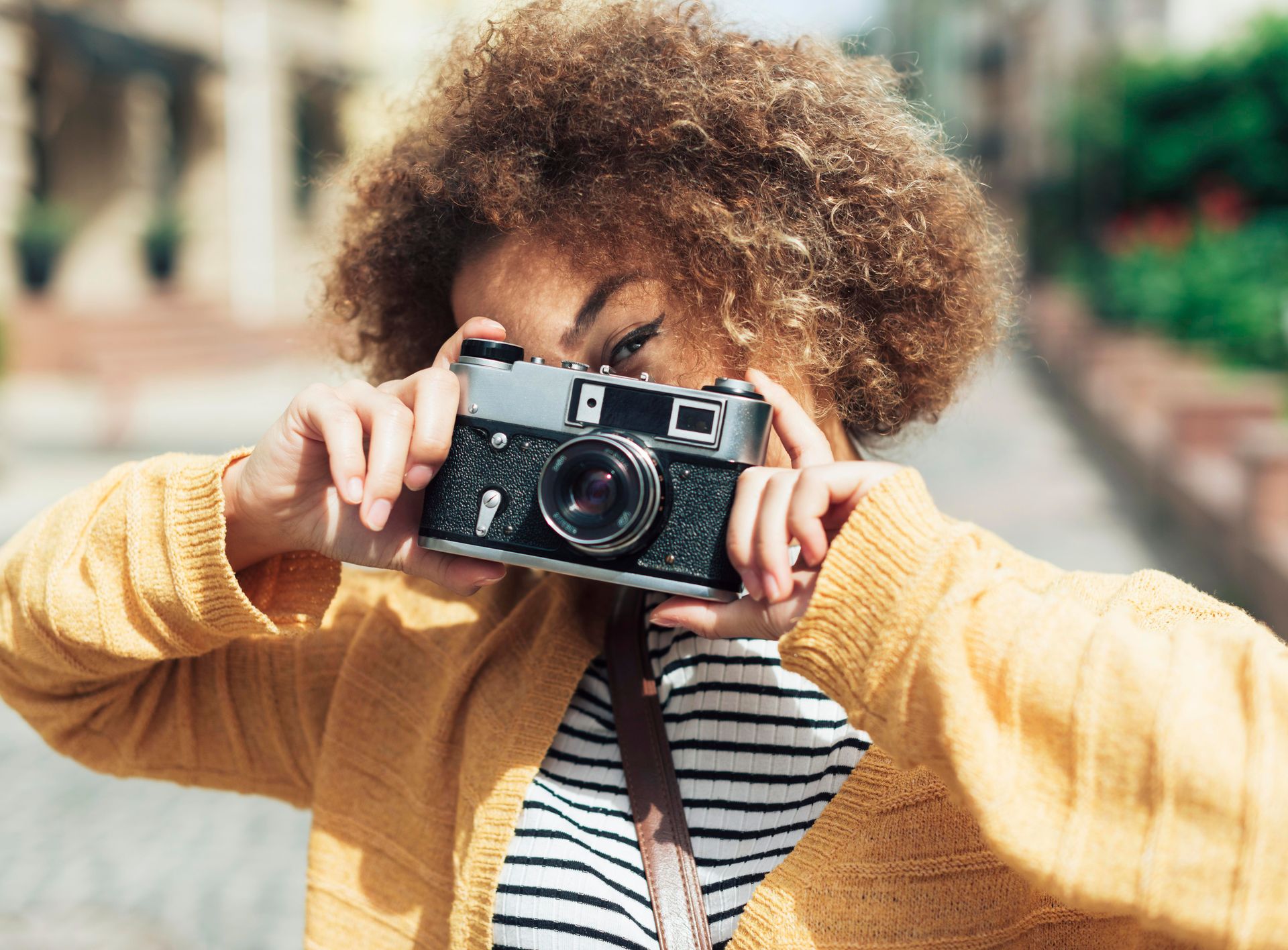 Une personne aux cheveux bouclés tient un appareil photo argentique vintage, vêtue d'un pull jaune sur un haut rayé. Le décor, une rue urbaine en plein air, évoque la créativité. Une personne aux cheveux bouclés tient un appareil photo argentique vintage, vêtue d'un pull jaune sur un haut rayé. Le décor, une rue urbaine en plein air, évoque la créativité.