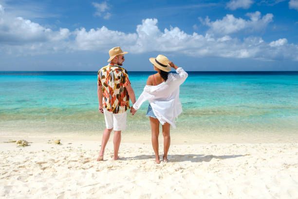 Couple holding hands on a white sand beach, looking out at clear turquoise ocean under a bright blue sky.