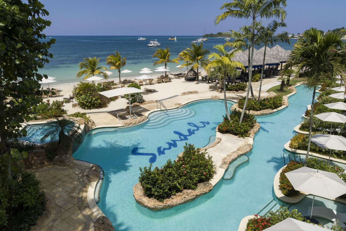 Luxury beachfront Sandals resort in Negril, Jamaica with winding turquoise pools, white sand beach, palm trees, and ocean views under a sunny blue sky.