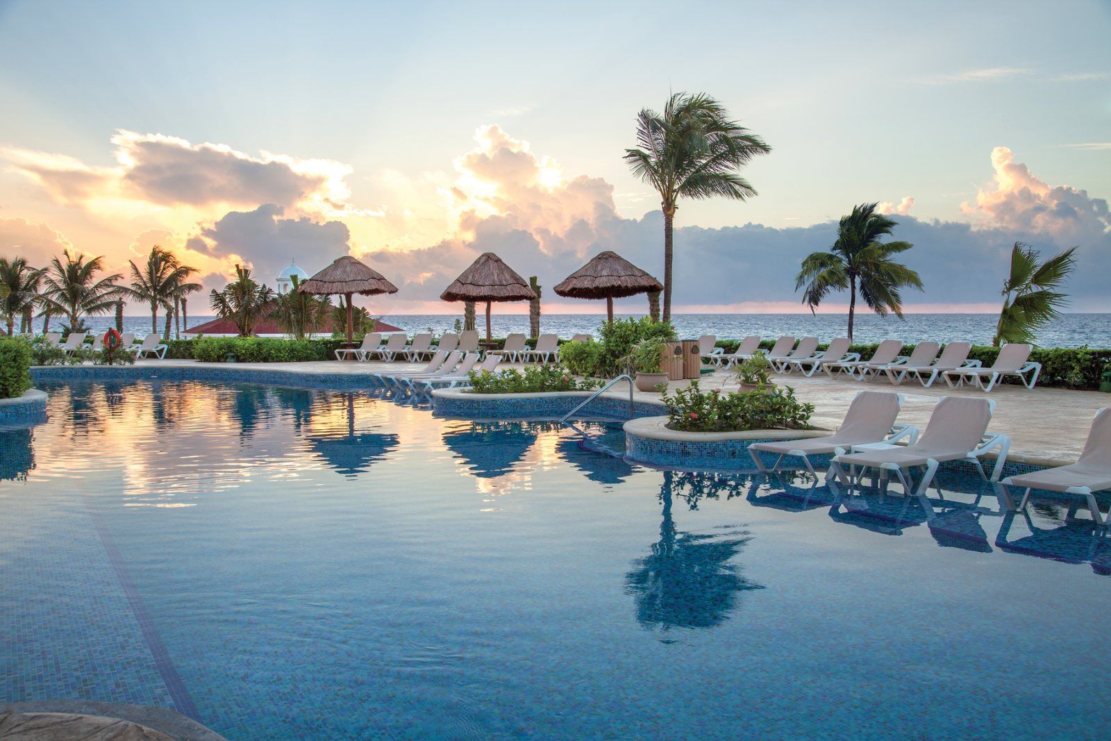 A peaceful tropical resort pool at sunrise with lounge chairs, palm trees, and thatched cabanas overlooking the ocean, reflecting the soft golden morning light. A peaceful tropical resort pool at sunrise with lounge chairs, palm trees, and thatched cabanas overlooking the ocean, reflecting the soft golden morning light.