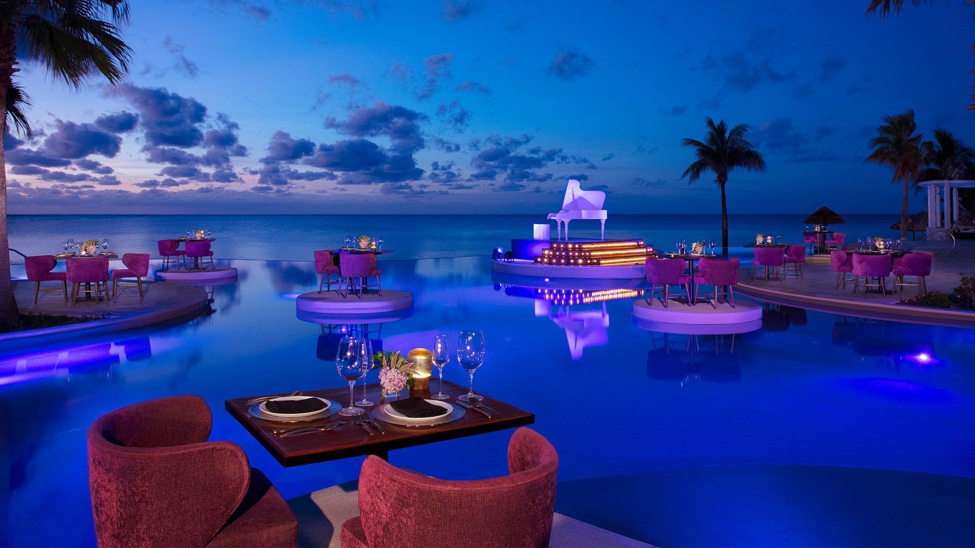 Romantic poolside dining setup at a luxury resort overlooking the ocean at twilight, with glowing lights and elegant table settings.
