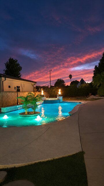 Illuminated pool, raised spa, palm island and fire bowls under a sunset sky