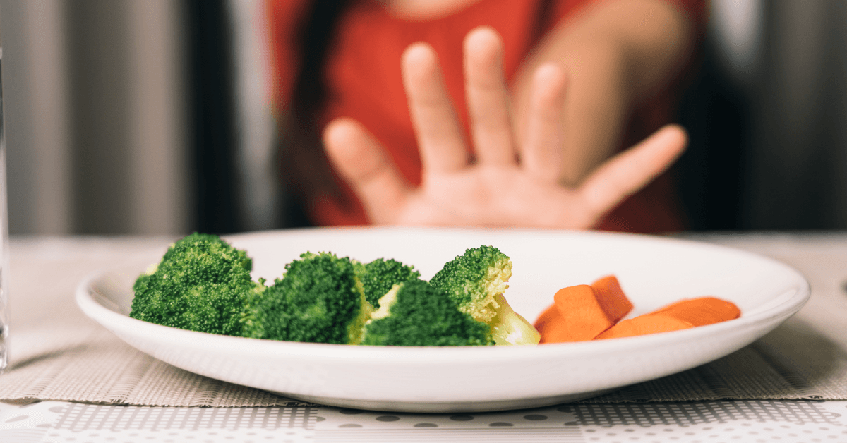 A hand held up in refusal in front of a plate of steamed broccoli and sliced carrots, symbolizing resistance to trying new things.