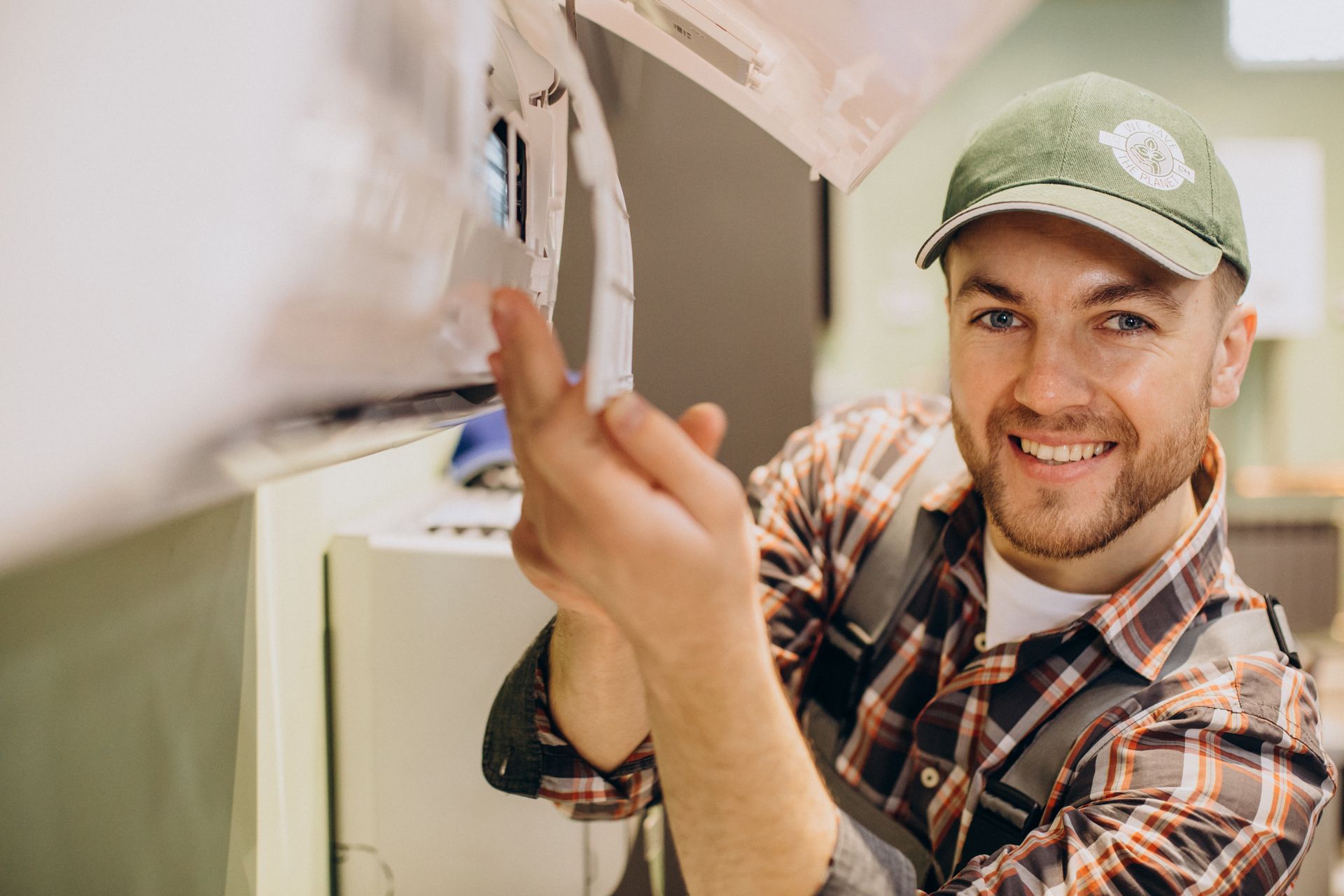 Un homme souriant, portant un chapeau vert et une chemise à carreaux, devant une climatisation. Un homme souriant, portant un chapeau vert et une chemise à carreaux, devant une climatisation.