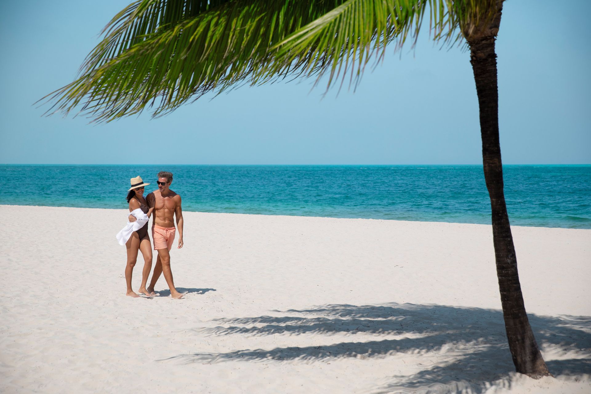 A couple walks barefoot along a bright white-sand beach beside turquoise ocean water, under a leaning palm tree on a sunny day. A couple walks barefoot along a bright white-sand beach beside turquoise ocean water, under a leaning palm tree on a sunny day.