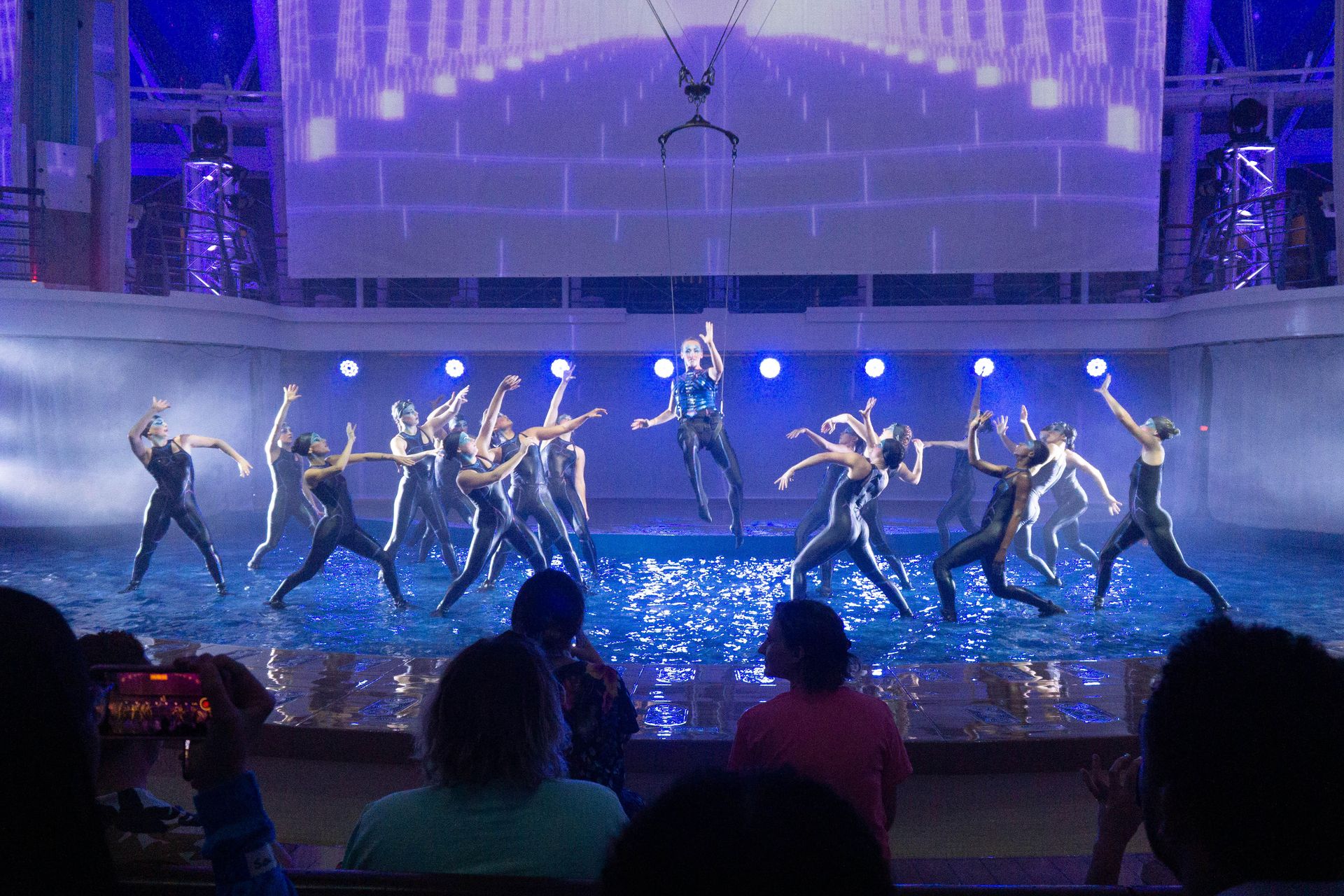 Acrobats perform a nighttime AquaTheater show on a cruise ship, dancing and leaping above a water stage as guests watch from poolside seating.