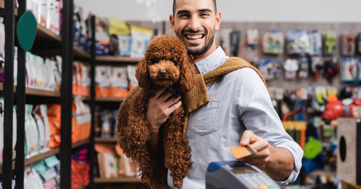 Close-up of a young man with facial hair smiling as he holds a brown poodle and pays at a pet store with a credit card. Close-up of a young man with facial hair smiling as he holds a brown poodle and pays at a pet store with a credit card.