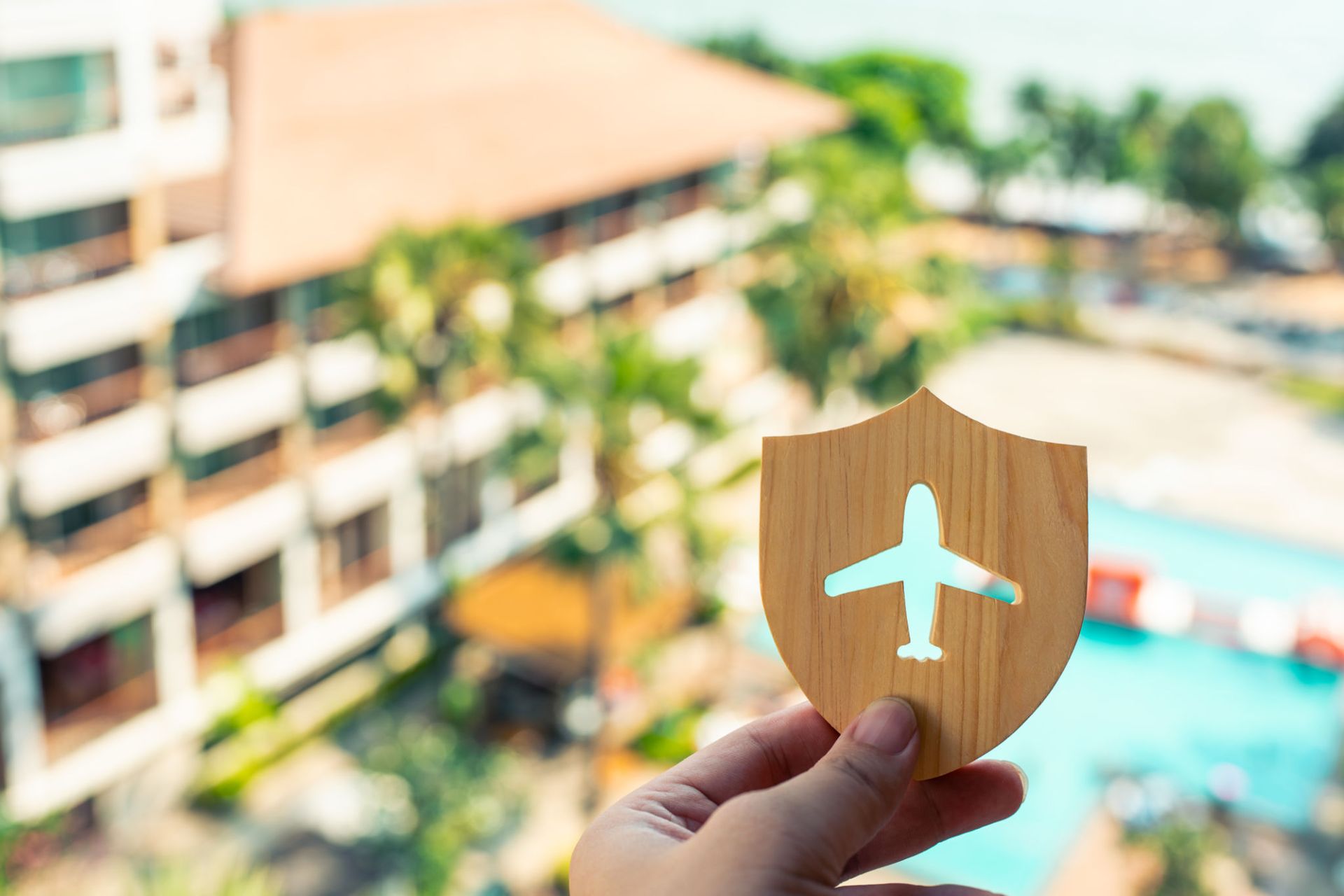 A hand holds a wooden shield cutout with an airplane symbol, symbolizing travel insurance, with a blurred background of a resort featuring palm trees and a swimming pool. A hand holds a wooden shield cutout with an airplane symbol, symbolizing travel insurance, with a blurred background of a resort featuring palm trees and a swimming pool.