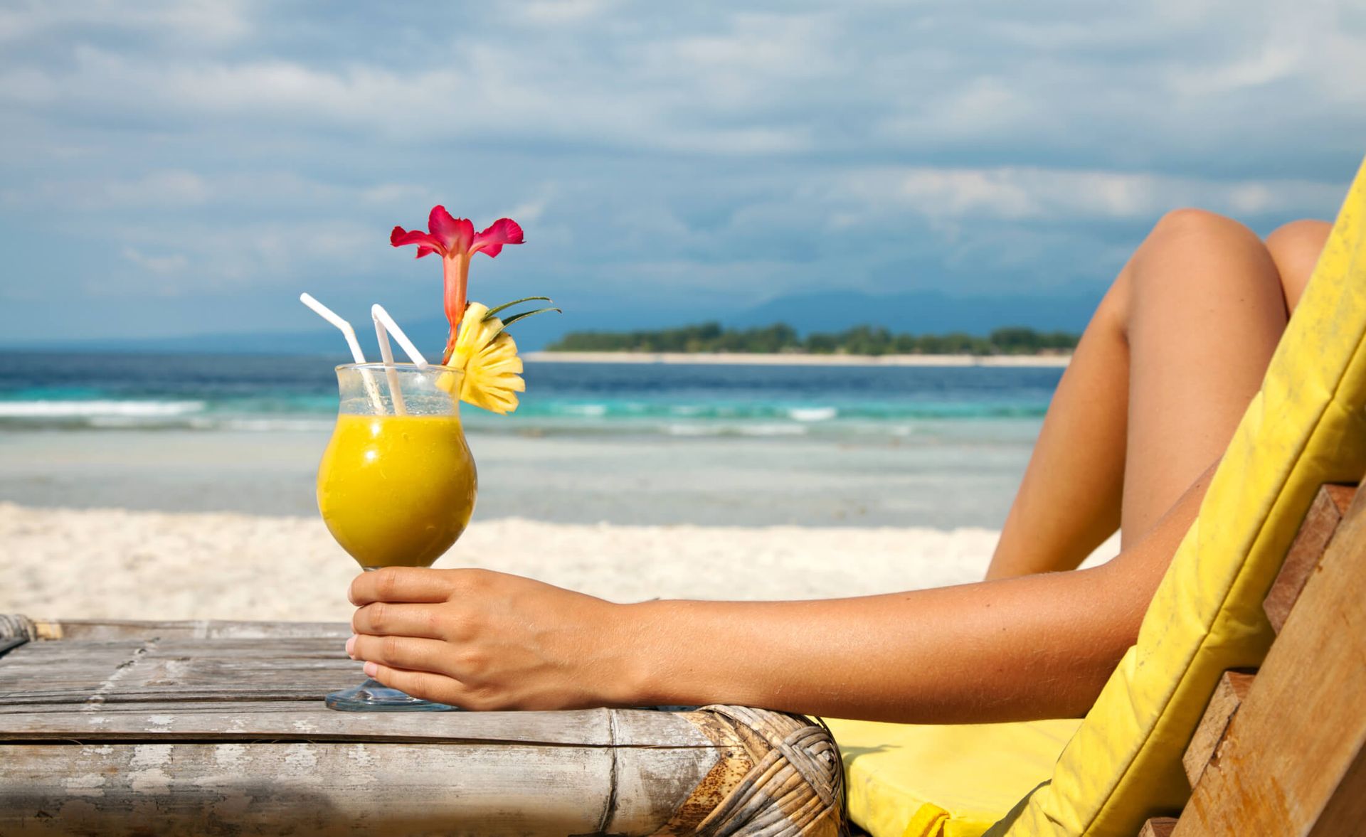 Person relaxing on a beach lounge chair holding a tropical drink garnished with flowers beside turquoise ocean waves. Person relaxing on a beach lounge chair holding a tropical drink garnished with flowers beside turquoise ocean waves.