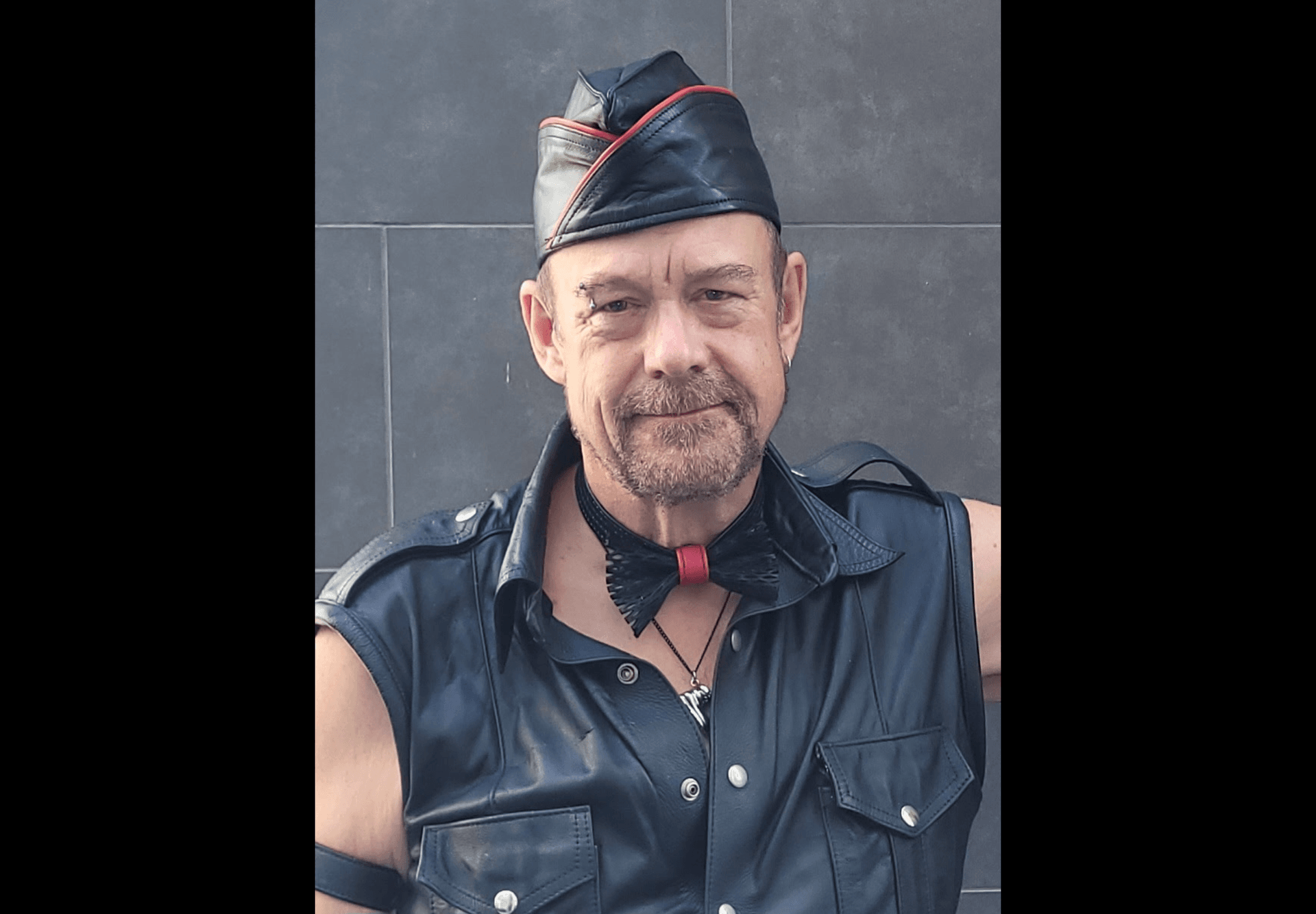 Scottie J in a black leather garrison cap with red trim and sleeveless leather vest looking directly at the camera against a dark stone wall Scottie J in a black leather garrison cap with red trim and sleeveless leather vest looking directly at the camera against a dark stone wall