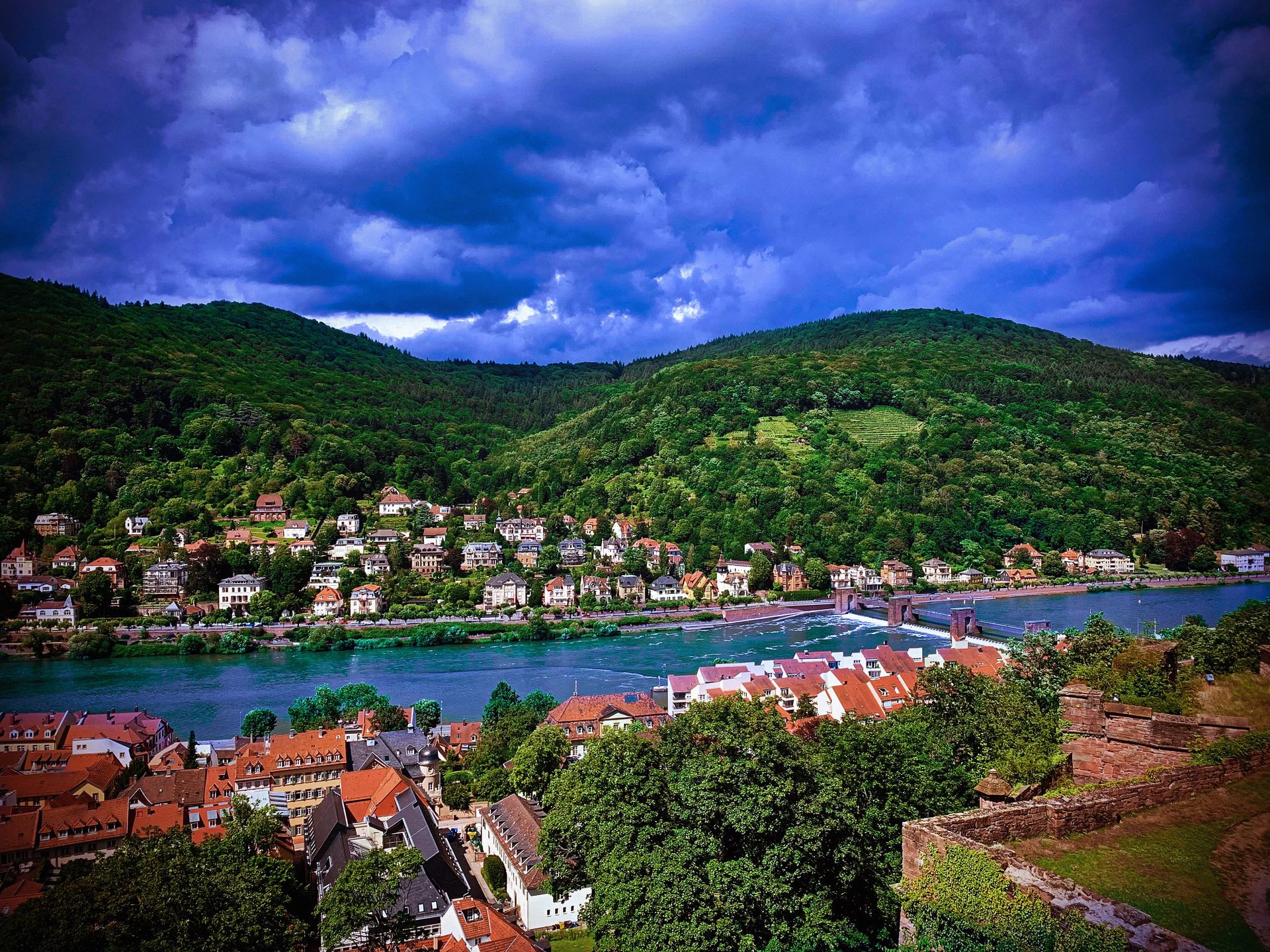A scenic view of a historic riverside town along the Rhine, with red-roofed houses, a stone bridge, lush green hills, and dramatic clouds over the landscape.