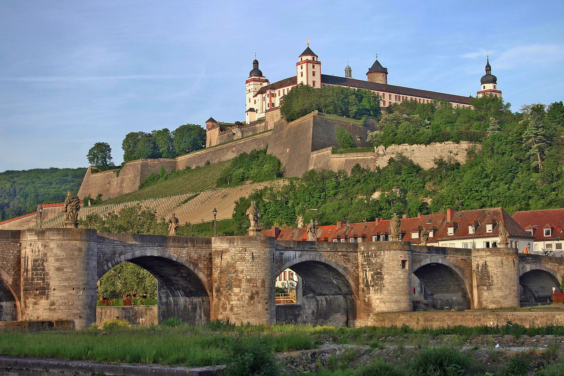 Historic stone bridge and Marienberg Fortress overlooking Würzburg, Germany, with vineyard-covered hillsides and traditional rooftops along the river.