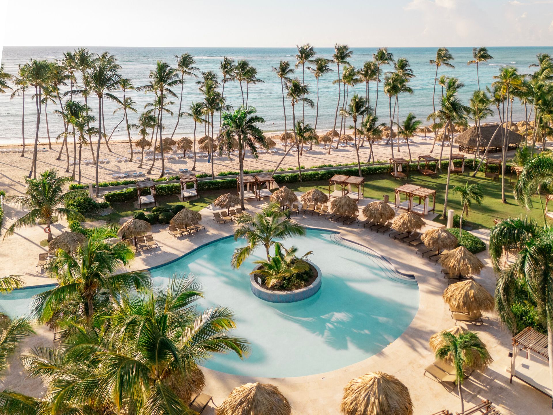 Aerial view of a tropical beachfront resort with a curved pool, palm trees, lounge chairs, and ocean waves along a sandy beach.