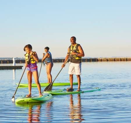 A family enjoying paddleboarding together on calm blue water, wearing life jackets and smiling under the warm sunlight. A family enjoying paddleboarding together on calm blue water, wearing life jackets and smiling under the warm sunlight.