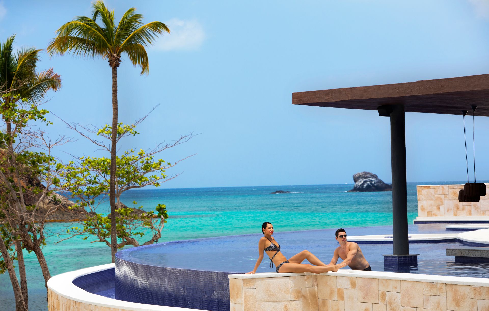 Couple relaxing by an infinity pool overlooking turquoise ocean water with palm trees and rocky shoreline in the background at a tropical resort. Couple relaxing by an infinity pool overlooking turquoise ocean water with palm trees and rocky shoreline in the background at a tropical resort.