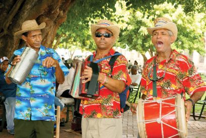 Three musicians in colorful tropical shirts and straw hats performing outdoors with drums, a guira, and an accordion under a large tree.