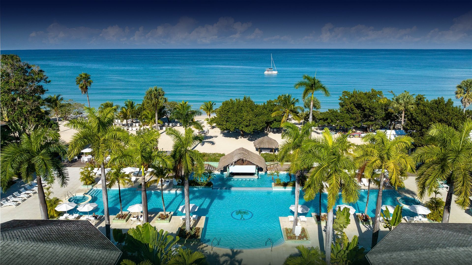 Luxury beachfront resort in Negril, Jamaica featuring a large turquoise pool, tropical palm trees, white sand beach, and calm Caribbean ocean views with a sailboat in the distance.