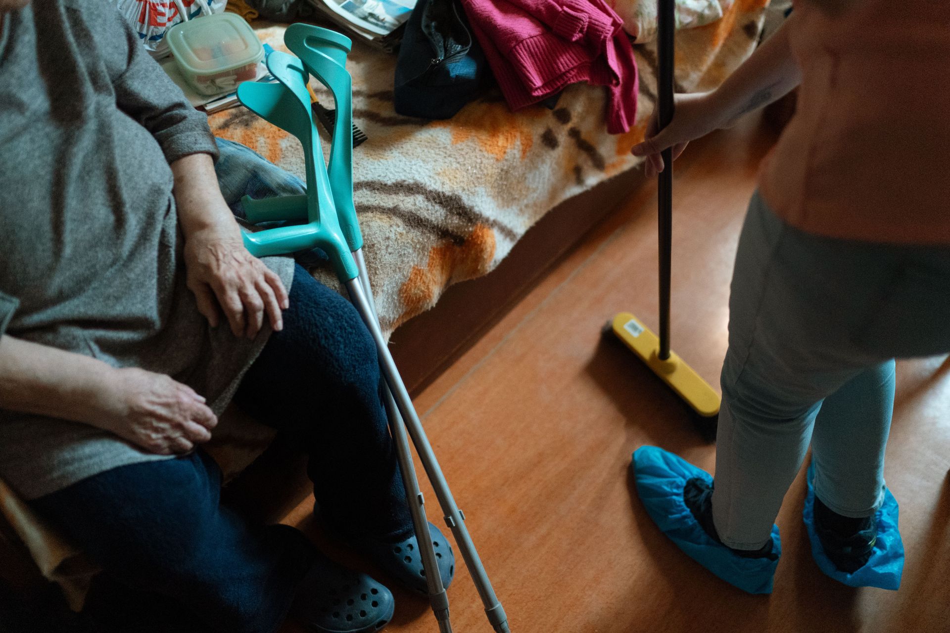 Close up of a faceless man with crutches and the legs of a woman in blue shoe covers sweeping the floor