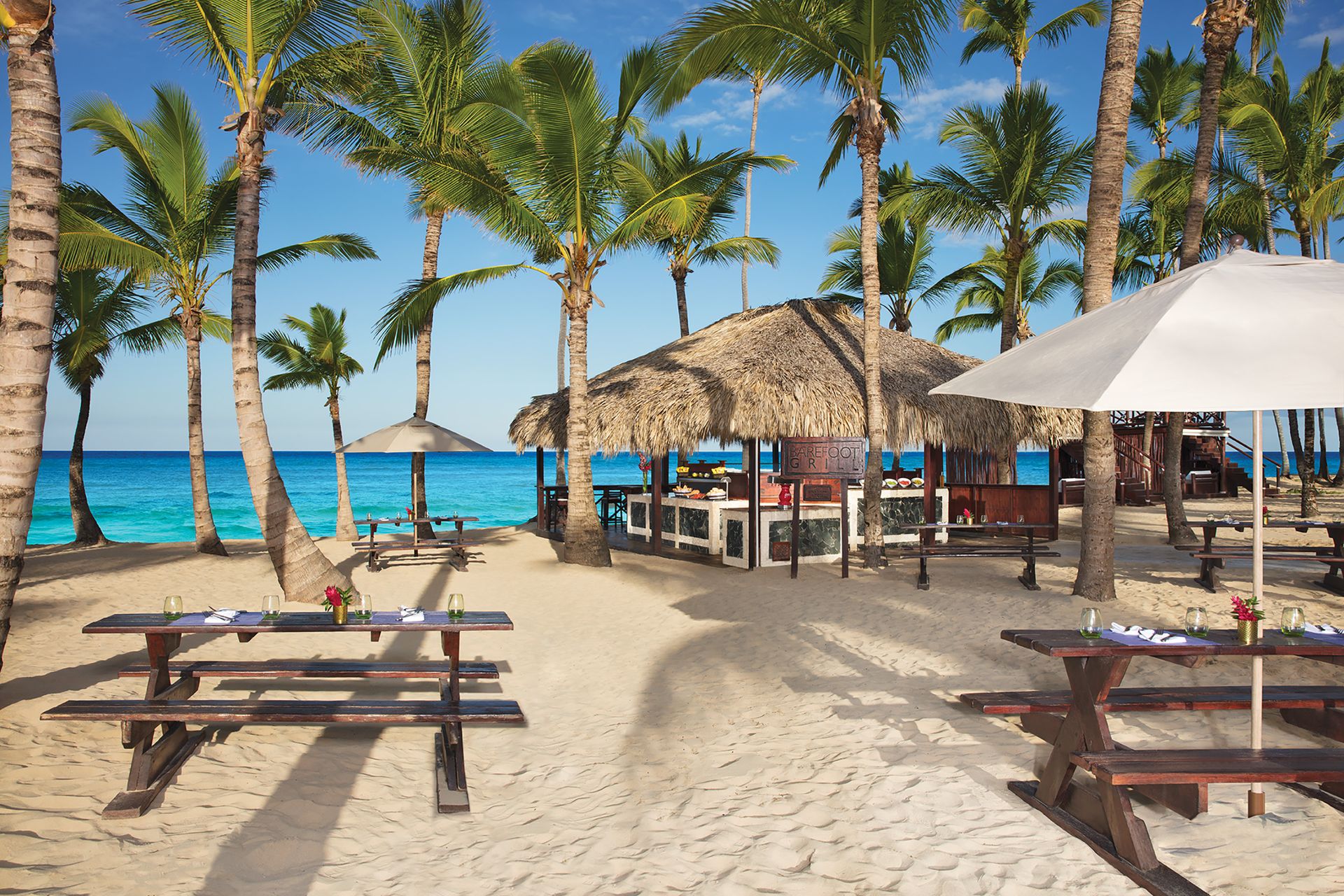 Beachfront restaurant with thatched roof and wooden picnic tables set on soft sand surrounded by palm trees, overlooking turquoise ocean water under a clear blue sky. Beachfront restaurant with thatched roof and wooden picnic tables set on soft sand surrounded by palm trees, overlooking turquoise ocean water under a clear blue sky.