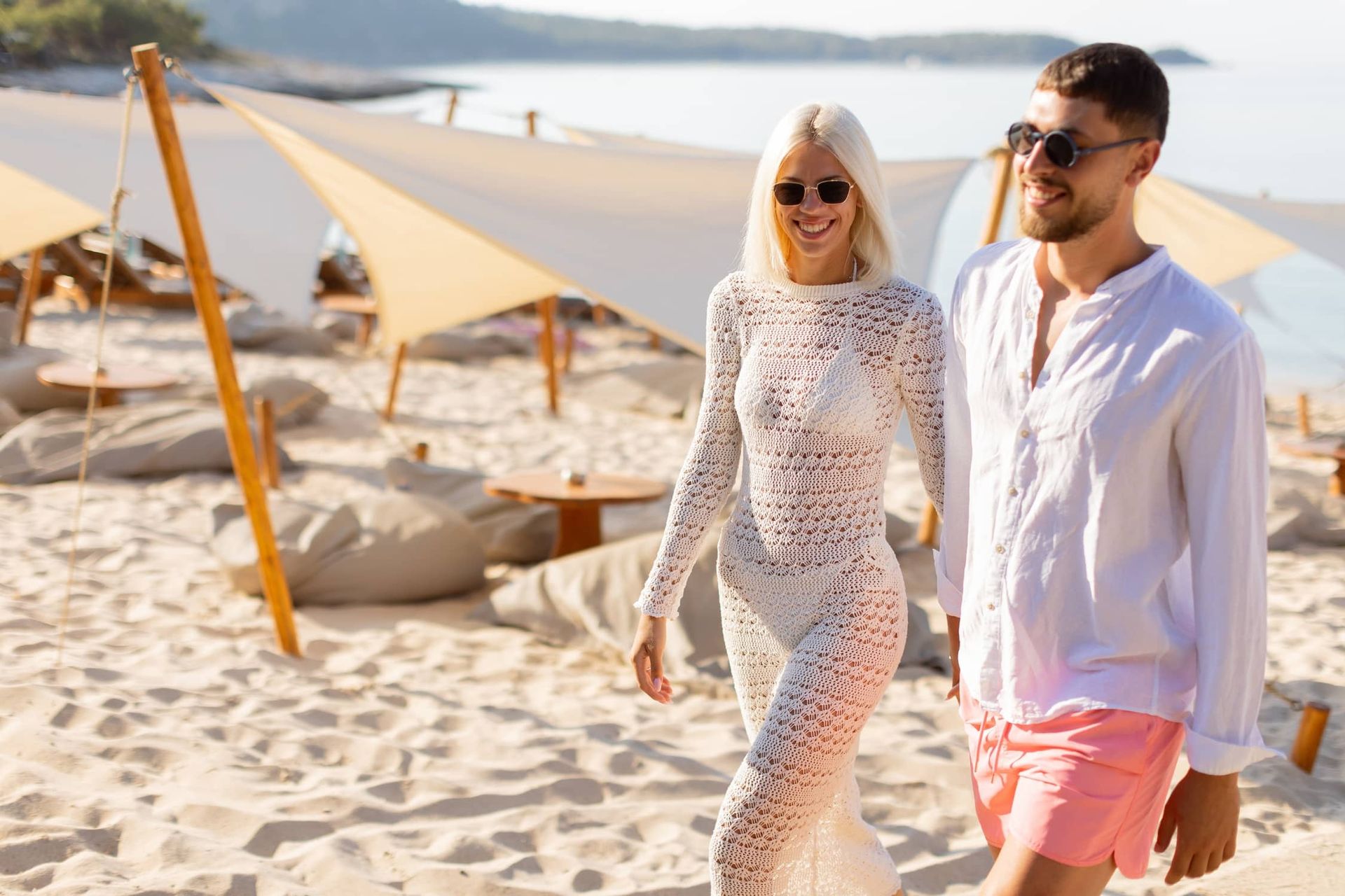 A smiling couple strolls along a white sand beach at a luxury resort, wearing stylish resort attire and sunglasses, with shaded lounge areas and calm ocean views in the background on a sunny day.
