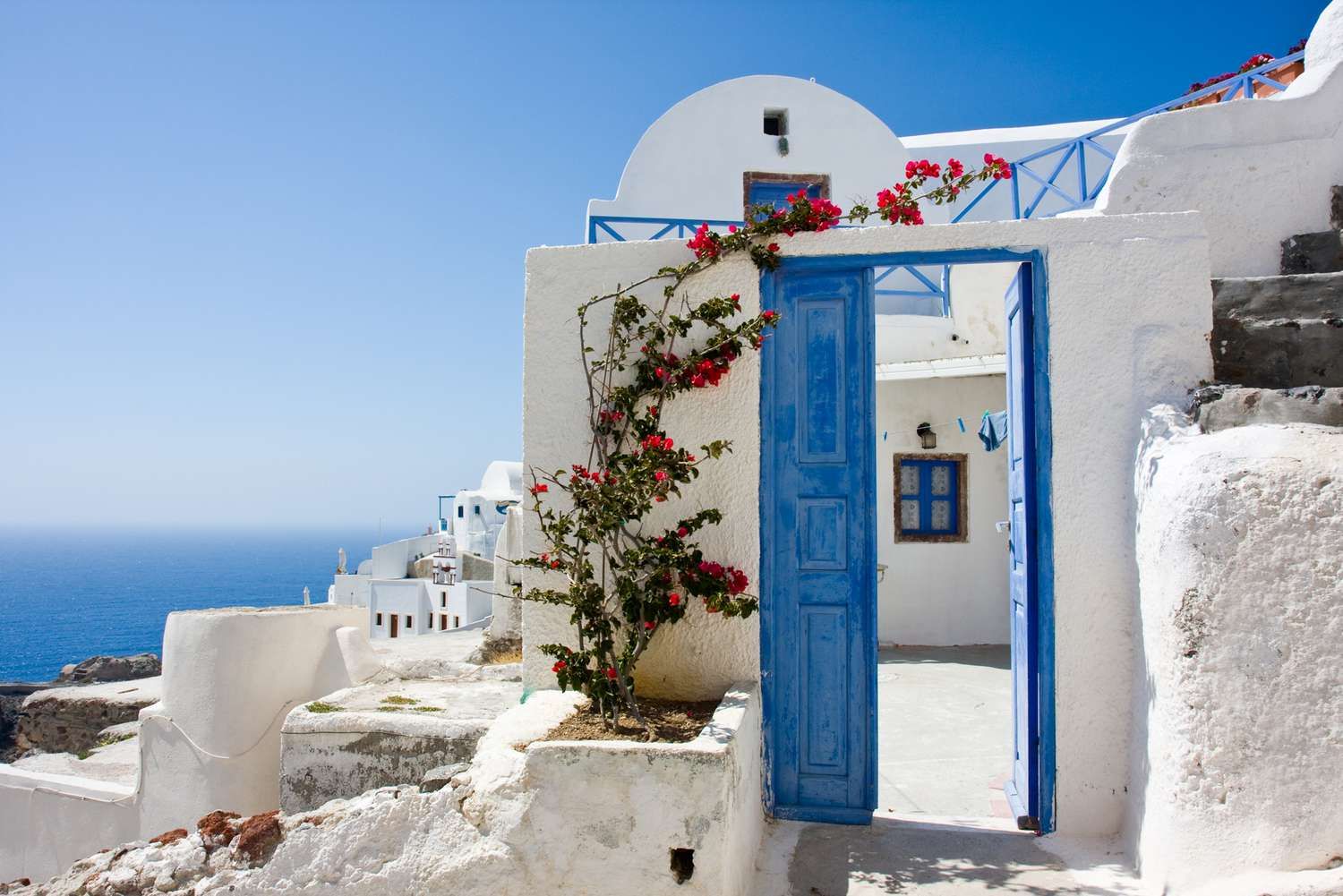 Whitewashed Santorini home with a bright blue doorway, blooming bougainvillea, and stunning views of the Aegean Sea under a clear blue sky.