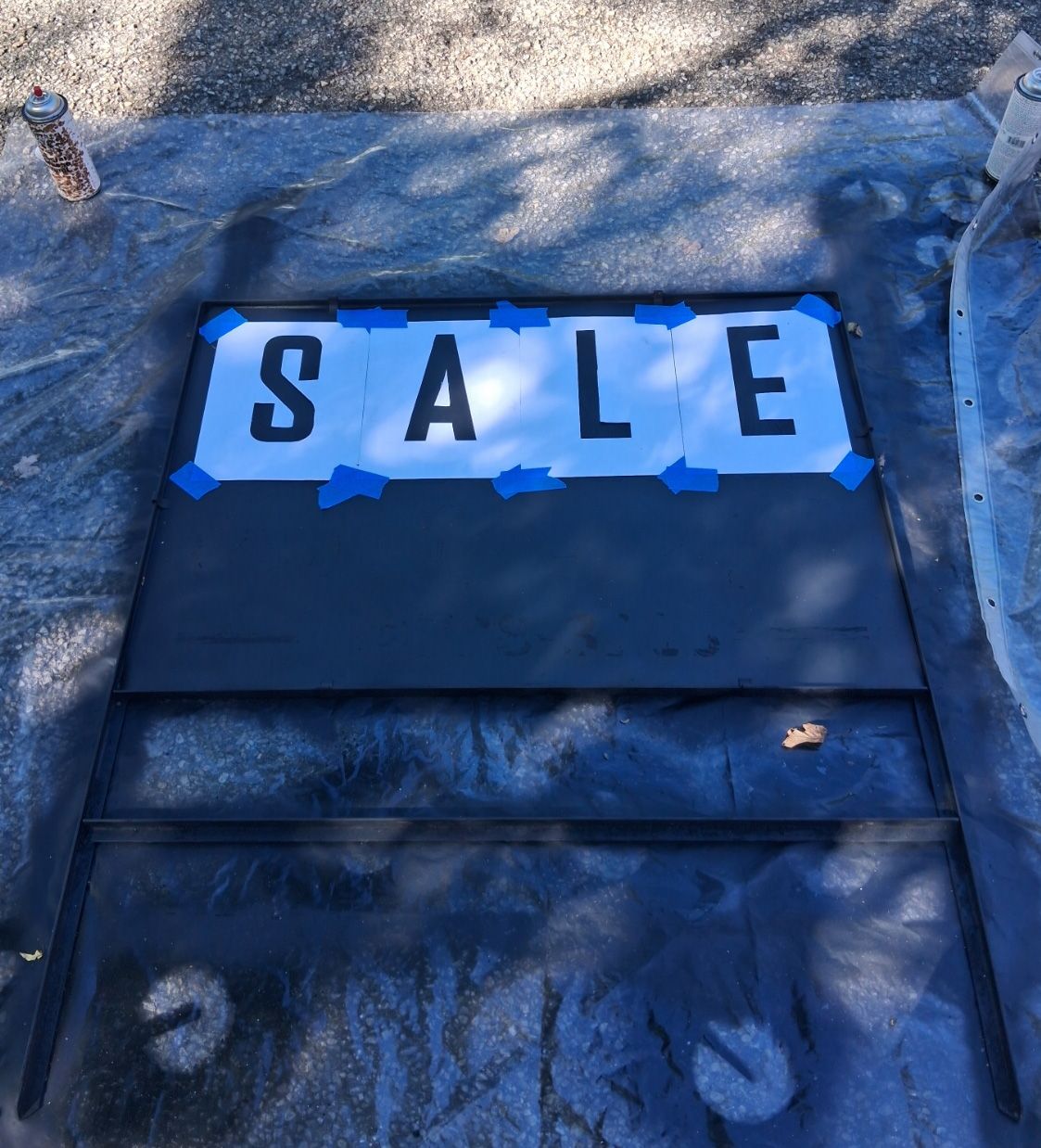 “Large DIY garage sale sign with oversized paper stencils spelling SALE taped to a black board before painting”