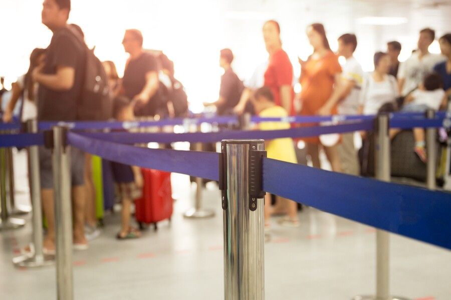 Travelers standing in a long airport security or check-in line with blue queue barriers and luggage visible in the background. Travelers standing in a long airport security or check-in line with blue queue barriers and luggage visible in the background.