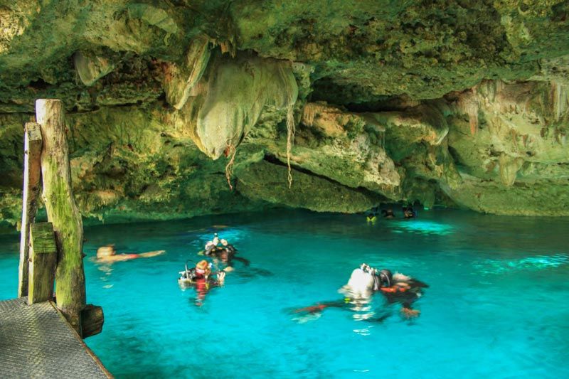 People swim and snorkel in a turquoise cenote inside a limestone cave with rock formations overhead.