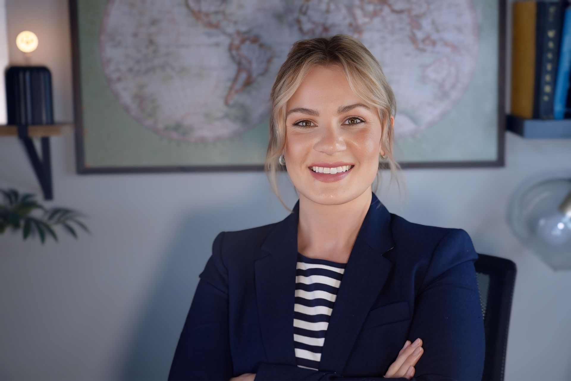 Smiling woman in a blazer with folded arms, standing in front of a world map.
