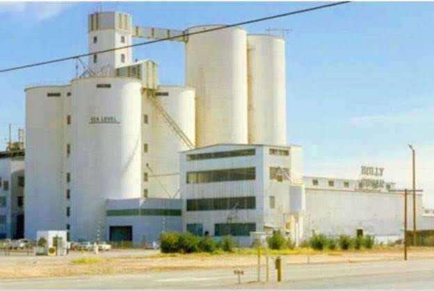 The image shows a large industrial building with tall white silos and adjoining structures, likely part of the Las Qoray Fish Factory. It appears to be a fish-processing or storage plant located in a dry, sunny area. The image shows a large industrial building with tall white silos and adjoining structures, likely part of the Las Qoray Fish Factory. It appears to be a fish-processing or storage plant located in a dry, sunny area.