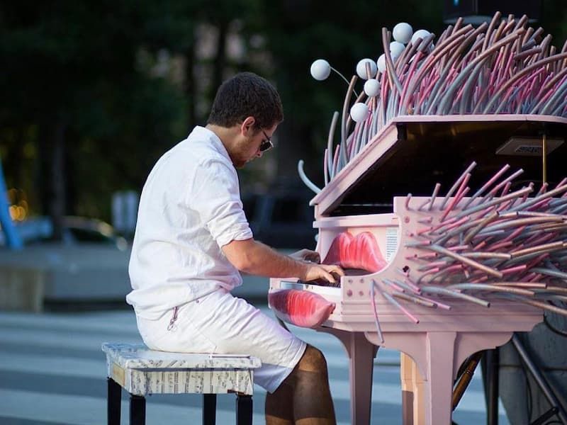 man with sunglasses plays grand piano with sculpted lips above and below the keys and bright pink and blue poles extruding from the body of the instrument