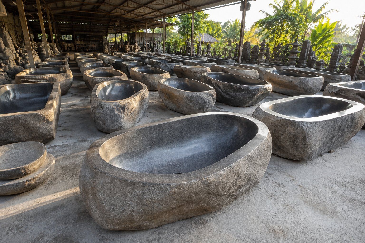 Rows of hand-carved river stone bathtubs in an open-air Bali stone yard, each a single hollowed boulder with a raw natural exterior and a smooth polished dark interior. Rows of hand-carved river stone bathtubs in an open-air Bali stone yard, each a single hollowed boulder with a raw natural exterior and a smooth polished dark interior.