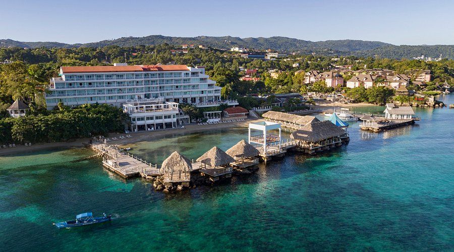 Aerial view of Sandals Ochi Beach Resort in Jamaica featuring beachfront buildings, overwater structures, and clear turquoise Caribbean waters.