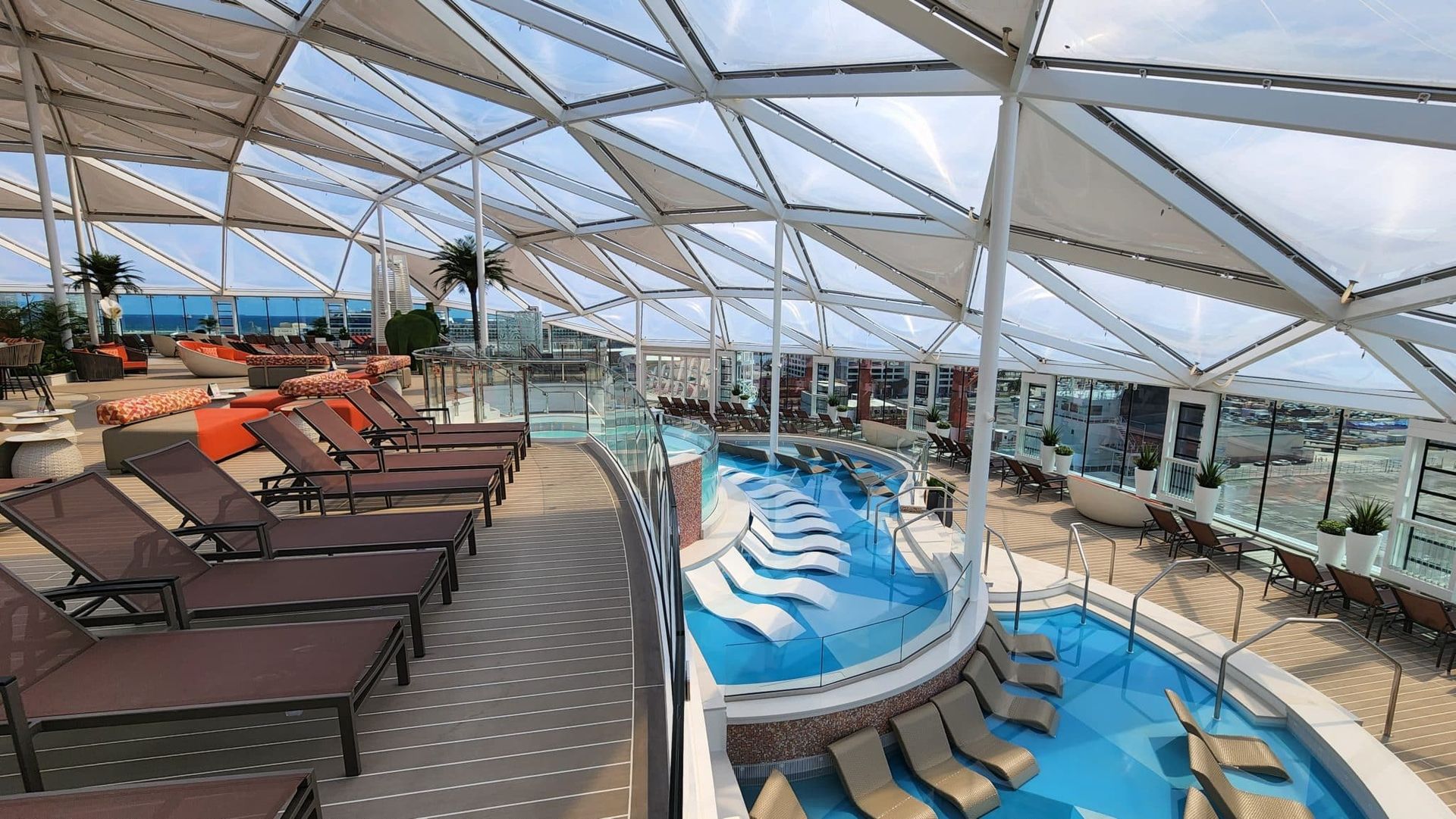 Covered cruise ship solarium featuring lounge chairs, pools, and ocean views under a glass roof
