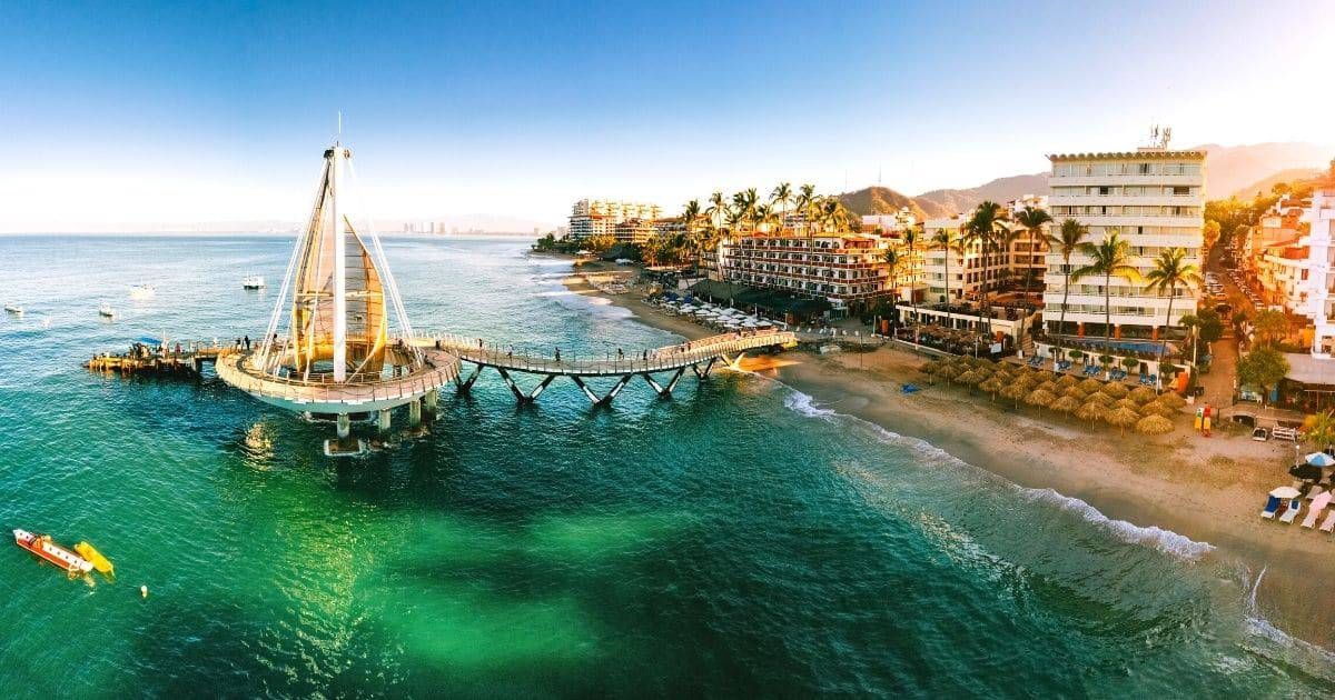 Aerial view of Puerto Vallarta's iconic pier extending into turquoise waters, surrounded by beachfront resorts, palm-lined shores, and scenic mountains. The destination showcases authentic Mexican charm, walkability, and a vibrant coastal atmosphere.