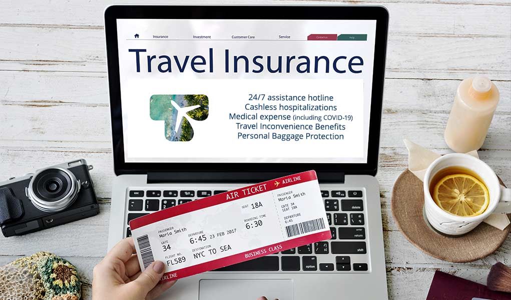 Hand holding airline ticket beside laptop showing “Travel Insurance” on a desk with camera and a drink.