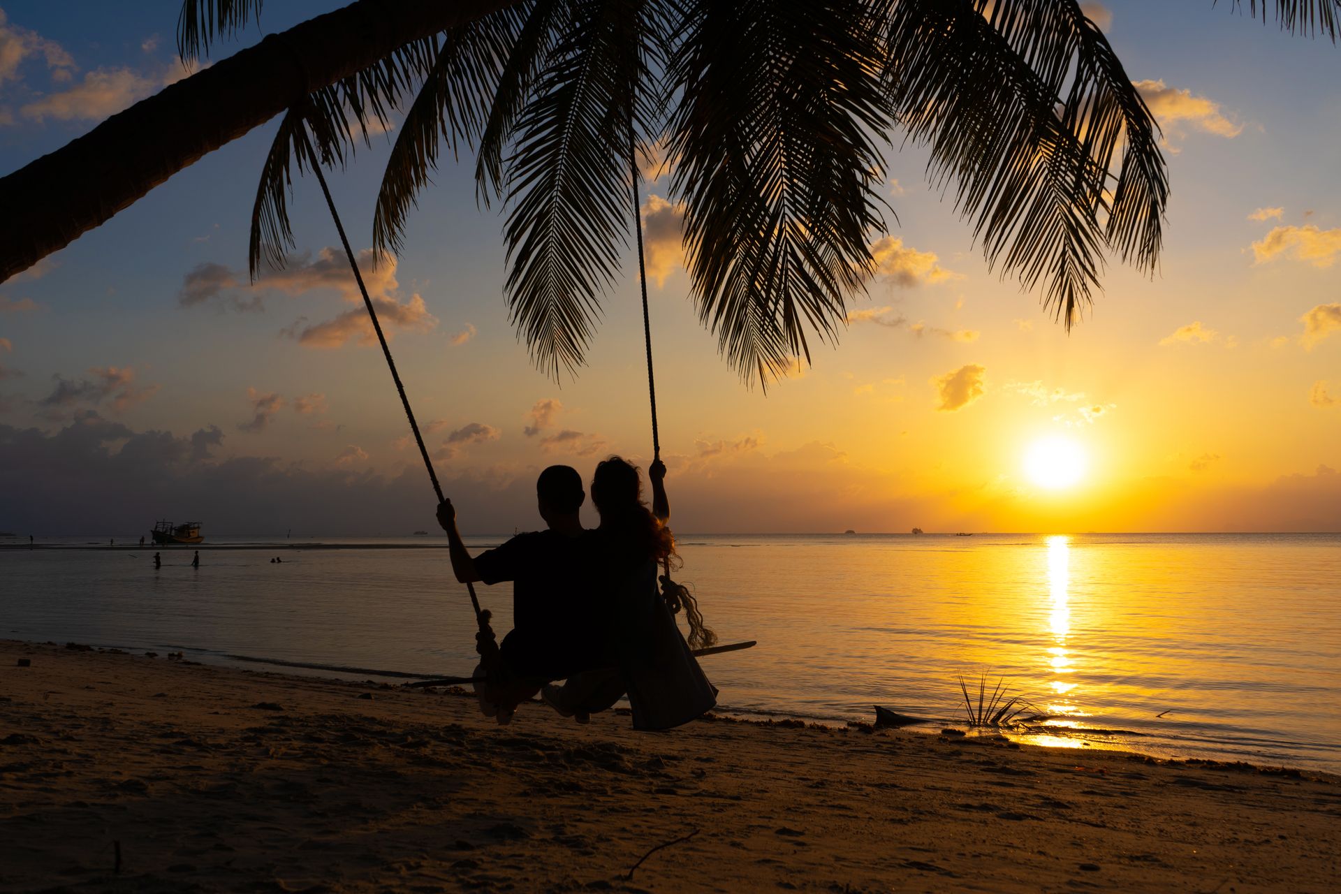 Silhouette of a couple sitting on a swing tied to a palm tree, watching a golden sunset over a calm tropical ocean. Silhouette of a couple sitting on a swing tied to a palm tree, watching a golden sunset over a calm tropical ocean.