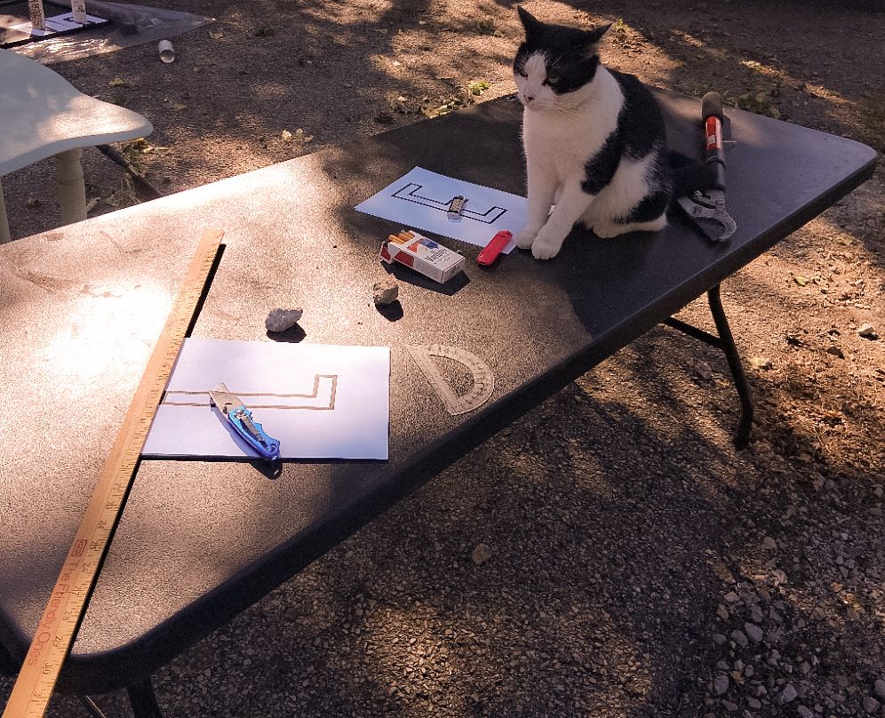 “Black-and-white cat sitting on a work table with paper stencils, ruler, and utility knife for a DIY garage sale sign project”