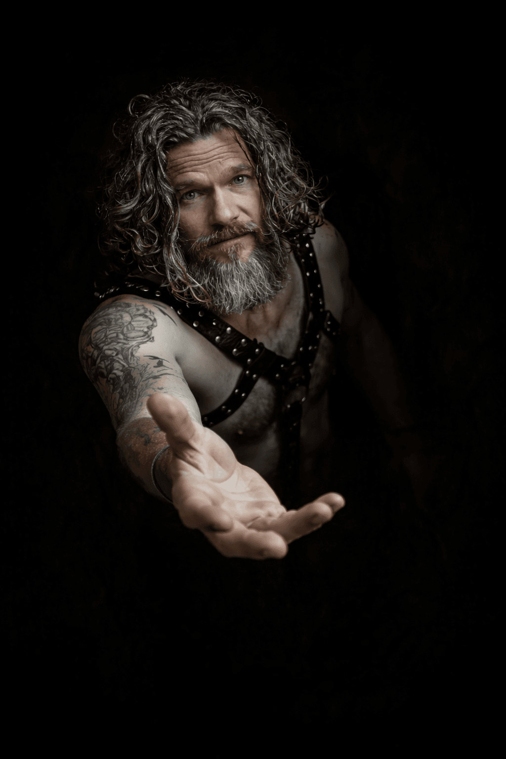 Sir Christopher in leather harness extending his hand forward against a dark background, symbolizing connection and guidance.