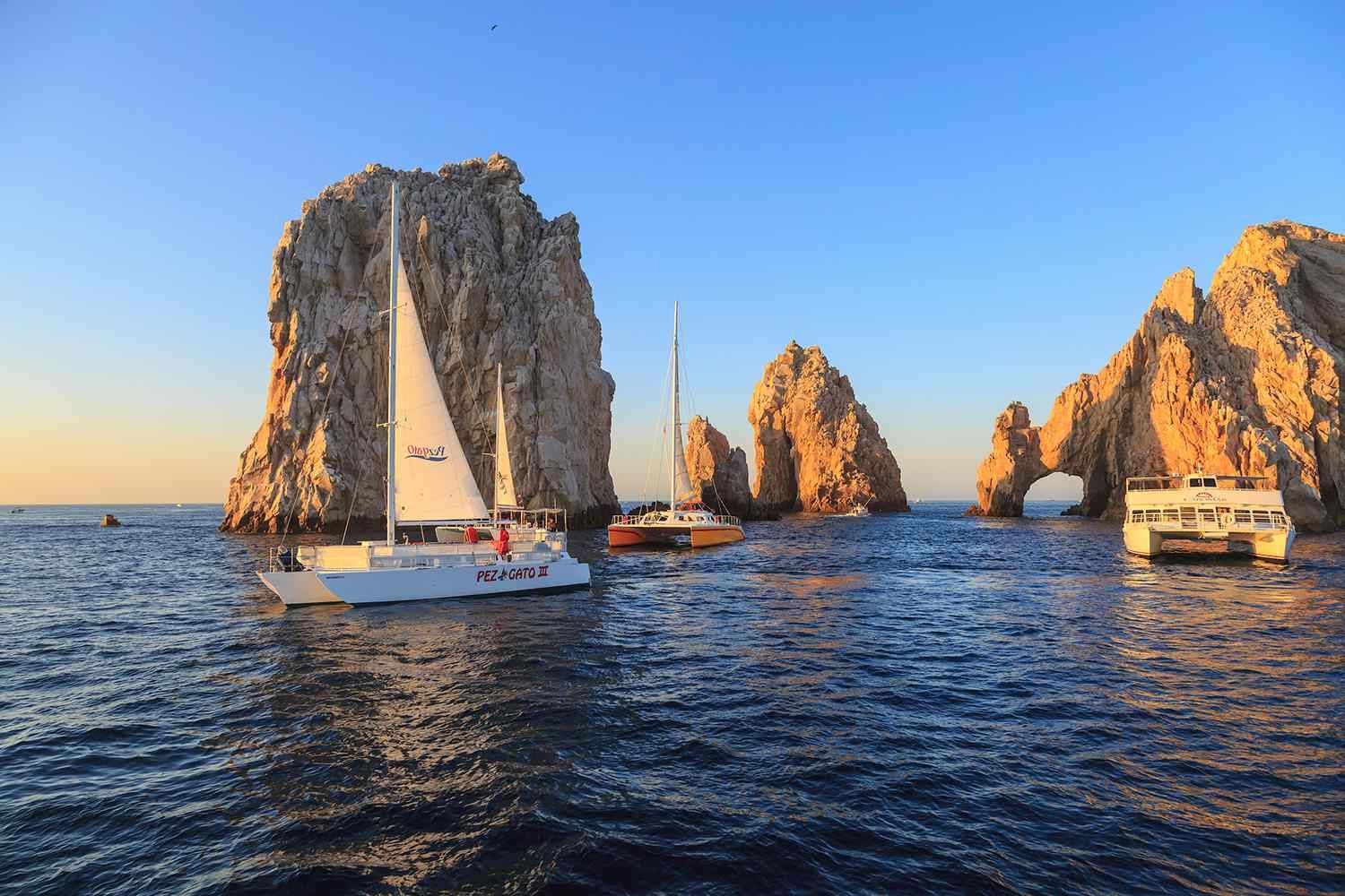 Sailboats and luxury catamarans glide across the waters of Los Cabos at sunset, framed by the iconic Arch rock formations. The scene captures the destination's blend of upscale adventure, wellness, and elevated coastal escapes.