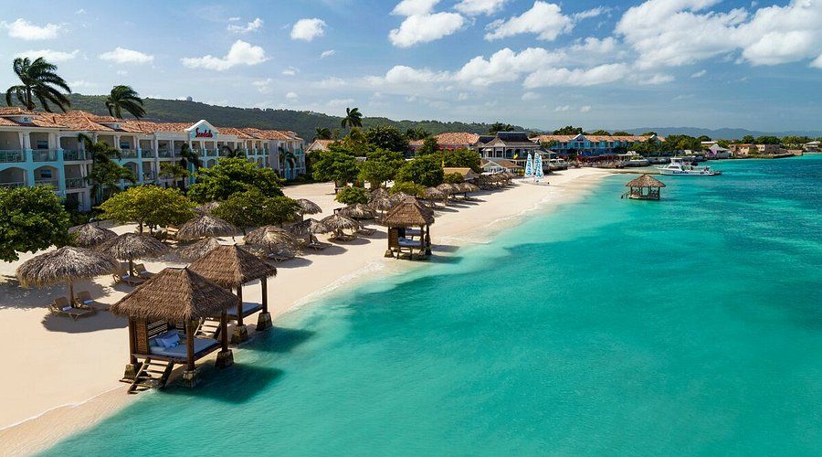 Beachfront view of Sandals Montego Bay resort with white sand, thatched cabanas, turquoise water, and tropical buildings along the shoreline in Jamaica.