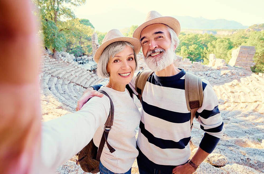 Smiling mature couple wearing hats and backpacks taking a selfie at ancient ruins during a sunny outdoor vacation.